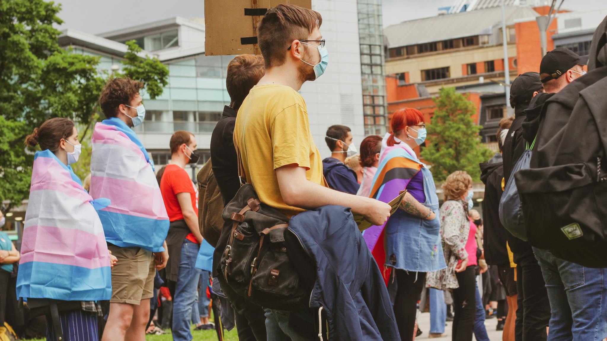 A diverse group of masked individuals gather outdoors supporting LGBTQ rights with pride flags.
